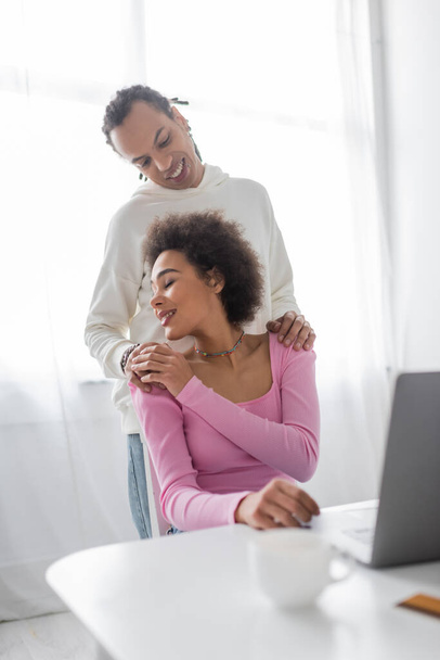 African american woman touching hand of boyfriend near laptop and coffee at home  - Photo, Image