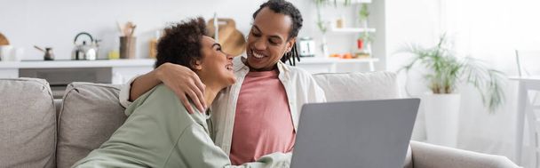 Smiling african american woman hugging boyfriend near laptop on couch at home,, banner  - Фото, изображение