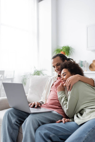 Sonriente pareja afroamericana abrazándose mientras usa la computadora portátil en casa  - Foto, imagen
