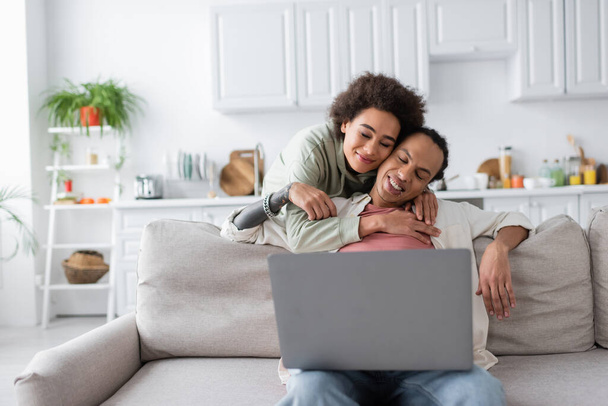 Young african american woman hugging boyfriend wit laptop at home  - Foto, Imagen