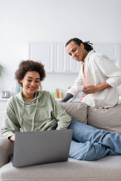 Cheerful african american woman using laptop near boyfriend with coffee at home  - Foto, Imagen