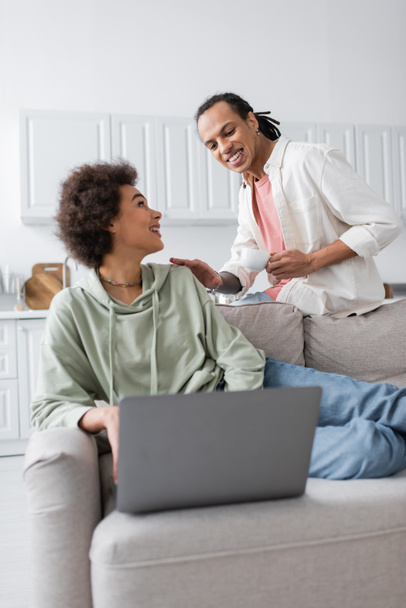Couple afro-américain positif avec café et ordinateur portable parlant à la maison  - Photo, image