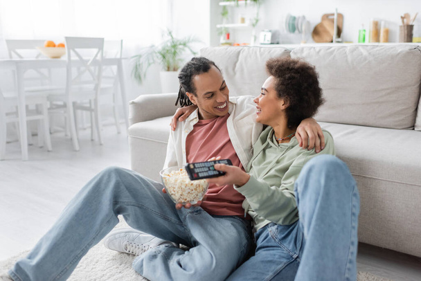 Positive african american couple with popcorn hugging while watching movie at home  - Fotografie, Obrázek