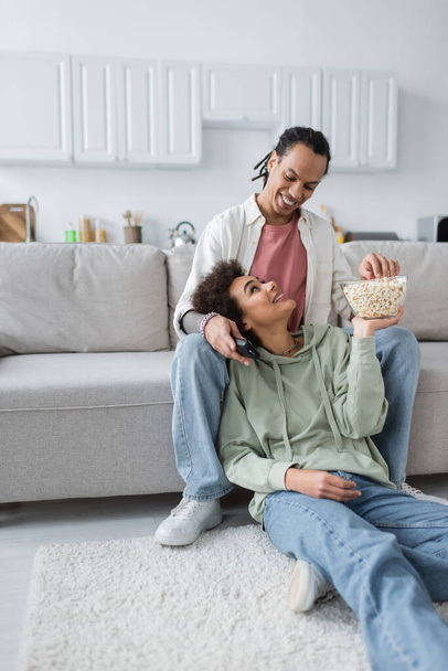 Sonriente mujer afroamericana sosteniendo palomitas de maíz cerca de novio con controlador remoto sentado en el sofá  - Foto, Imagen