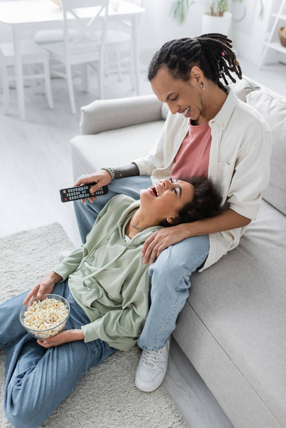 Positive african american couple holding remote controller and popcorn at home  - Fotoğraf, Görsel