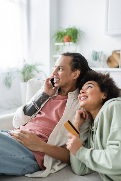 Kredi kartı tutan Afrikalı Amerikalı bir kadın ve evde akıllı telefondan konuşan erkek arkadaşına bakıyor. - Fotoğraf, Görsel