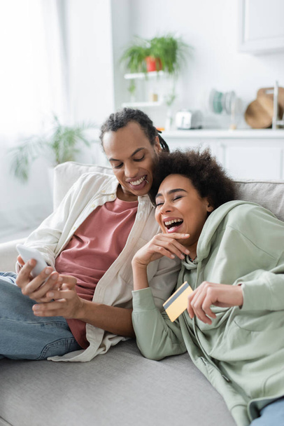 笑うアフリカ系アメリカ人女性保持クレジットカード近くボーイフレンドとともにスマートフォン上のソファ  - 写真・画像