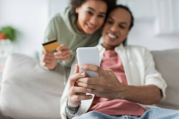 Pareja afroamericana borrosa usando teléfono inteligente y tarjeta de crédito durante las compras en línea en casa  - Foto, Imagen