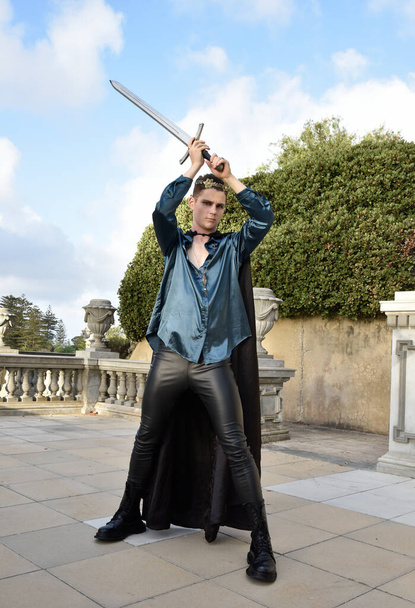  portrait of handsome brunette male model wearing fantasy medieval prince costume, romantic silk shirt, holding sword weapon.  historical castle location background with stone staircase. - Photo, Image