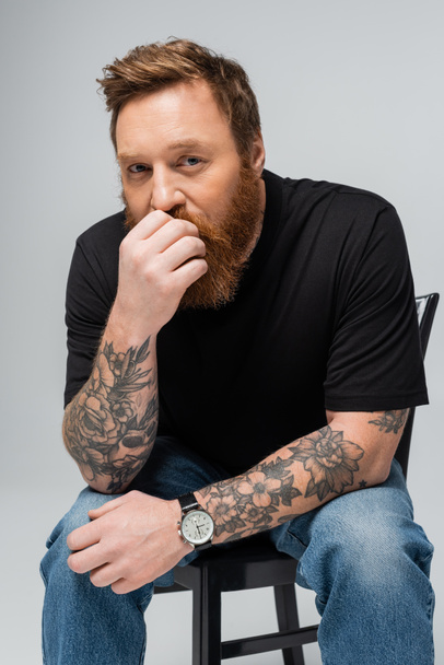 hombre tatuado pensativo en camiseta negra y reloj de pulsera sosteniendo la mano cerca de la cara y mirando a la cámara aislada en gris - Foto, imagen