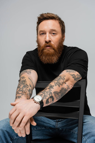 thoughtful bearded man in black t-shirt and wristwatch sitting on chair and looking at camera isolated on grey - Fotoğraf, Görsel