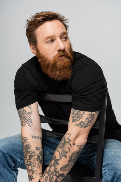 στοχαστικός άντρας με τατουάζ με μαύρο μπλουζάκι που κάθεται στην καρέκλα ενώ κοιτάζει μακριά, απομονωμένος στο γκρι - Φωτογραφία, εικόνα