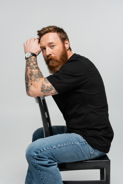 bearded and tattooed man in black t-shirt and jeans sitting on chair while looking at camera isolated on grey - Fotografie, Obrázek
