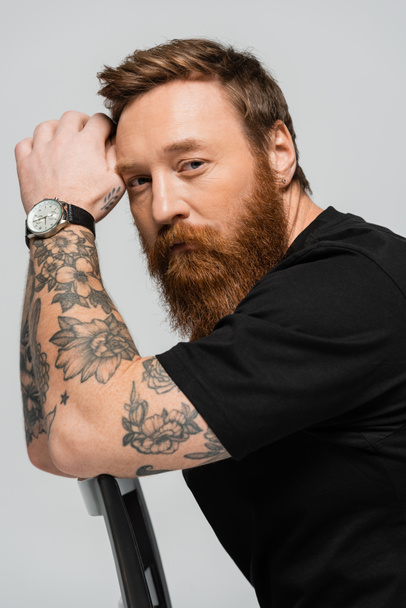 вдумливий татуйований чоловік в наручних годинниках і чорна футболка дивиться на камеру, сидячи ізольовано на сірому
 - Фото, зображення