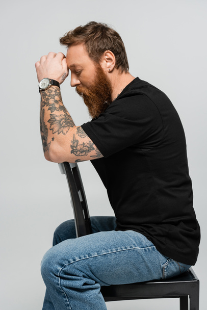 side view of tired bearded man in black t-shirt and wristwatch sitting with closed eyes and hands near head isolated on grey - Photo, Image