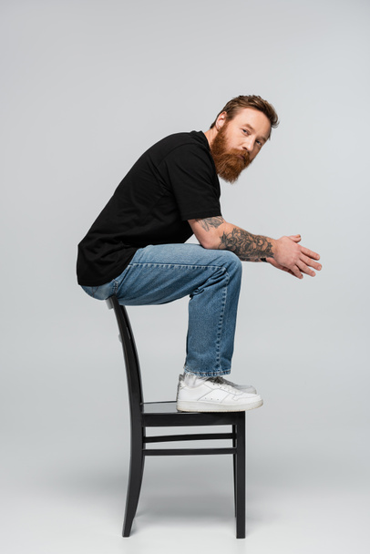 full length of bearded man in jeans sitting on chair back and looking at camera on grey background - Foto, immagini