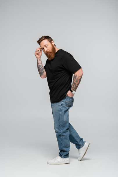 full length of thoughtful bearded man touching forehead and holding hand in back pocket of jeans on grey background - Photo, Image