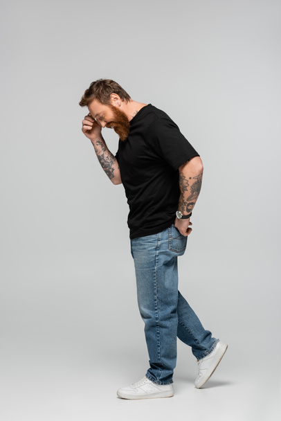 side view of smiling man in black t-shirt and jeans touching forehead while smiling on grey background - Zdjęcie, obraz