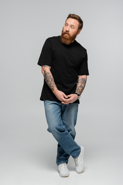 volledige lengte van getatoeëerde man met baard in jeans en zwart t-shirt staande met gebalde handen en wegkijkend op grijze achtergrond - Foto, afbeelding