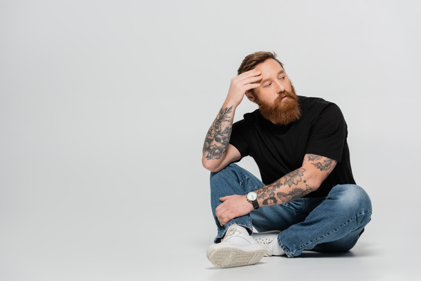 full length of thoughtful tattooed man in black t-shirt and jeans looking away while sitting on grey background - Foto, Imagem