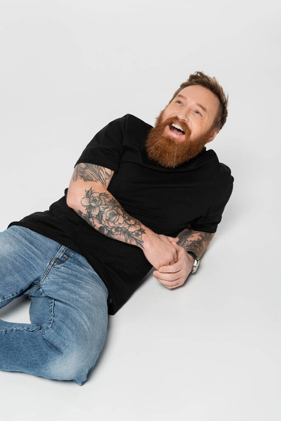 joyful bearded and tattooed man in jeans and black t-shirt laughing while lying on grey background - Foto, immagini