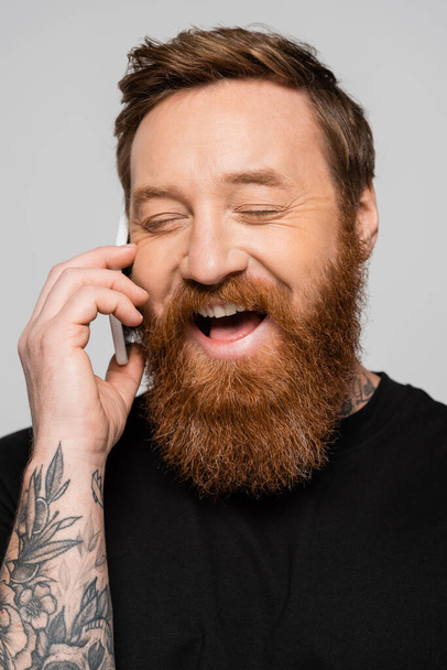 excited bearded man with closed eyes talking on mobile phone and laughing isolated on grey - Valokuva, kuva