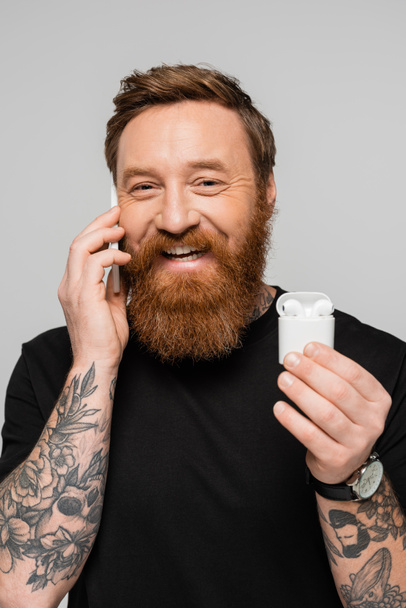 smiling tattooed man holding case with wireless earphones while talking on cellphone isolated on grey - Foto, Bild