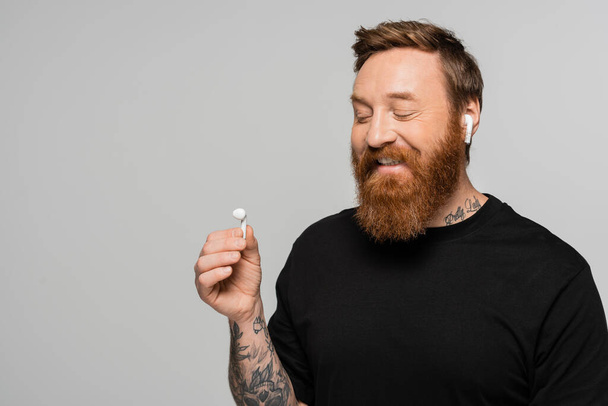 positive bearded man in black t-shirt looking at wireless earphone isolated on grey - Photo, Image