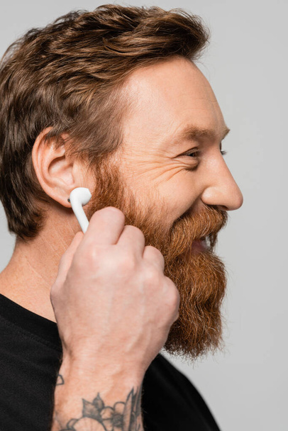 Вид сбоку жизнерадостного бородатого мужчины, держащего у уха беспроводной наушник, изолированный на сером фоне - Фото, изображение