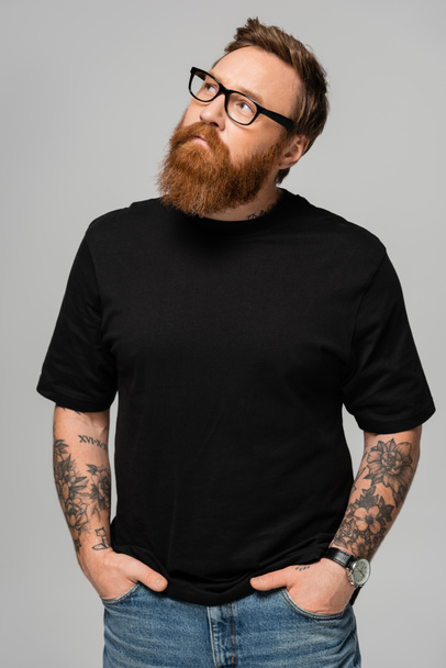 stylish bearded man in eyeglasses standing with hands in pockets of jeans and looking away isolated on grey - Photo, image