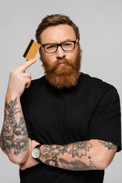 hombre tatuado reflexivo en gafas con tarjeta de crédito cerca de la cabeza mientras mira la cámara aislada en gris - Foto, imagen