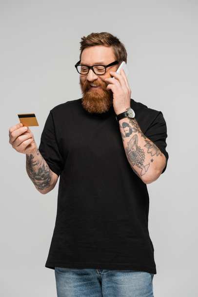 happy tattooed man in eyeglasses talking on mobile phone while holding credit card isolated on grey - Fotografie, Obrázek