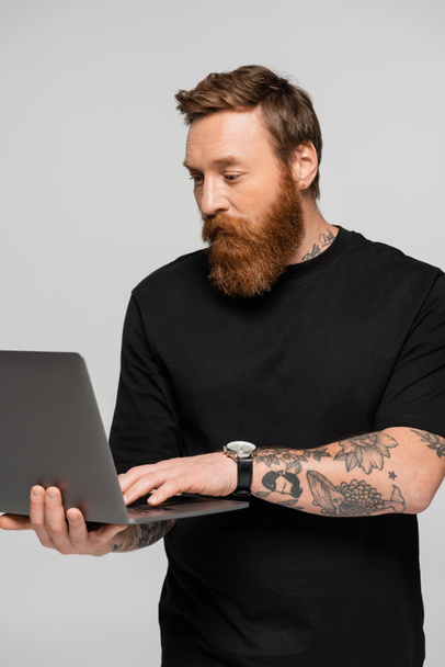 бородатый татуированный фрилансер в черной футболке с помощью ноутбука, изолированного на сером - Фото, изображение