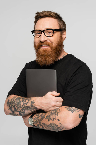 tyytyväinen parrakas freelancer silmälaseissa pitämällä kannettavaa tietokonetta ja hymyilemällä kameralle eristettynä harmaalla - Valokuva, kuva