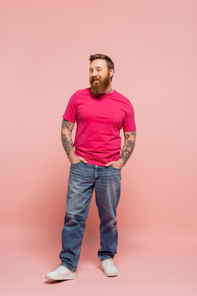 full length of happy bearded man in magenta t-shirt and jeans posing with hands in pockets on pink background - Foto, Bild