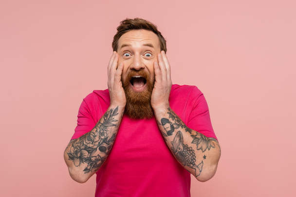 amazed bearded man in bright t-shirt touching face while looking at camera isolated on pink - Valokuva, kuva