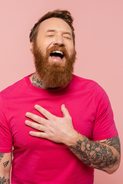 веселий бородатий чоловік торкається грудей і сміється із закритими очима ізольовано на рожевому
 - Фото, зображення