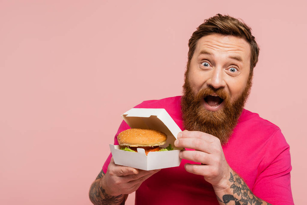 збуджений бородатий чоловік тримає картонну упаковку з бургер, дивлячись на камеру ізольовано на рожевому
 - Фото, зображення