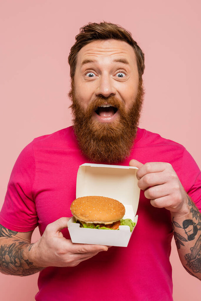 astonished tattooed man in bright t-shirt showing carton pack with delicious hamburger isolated on pink - Zdjęcie, obraz