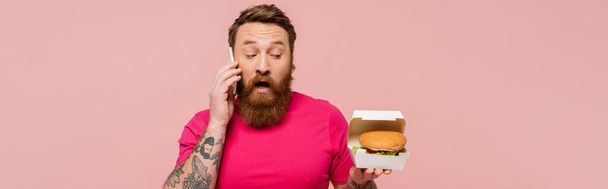 Aufgeregter Mann hält Kartonpackung mit leckerem Burger in der Hand, während er auf dem Smartphone mit rosa Banner spricht - Foto, Bild