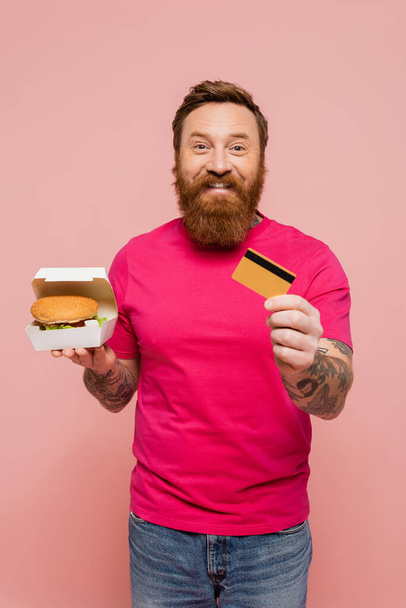 cheerful bearded man holding hamburger and showing credit card isolated on pink - Photo, Image