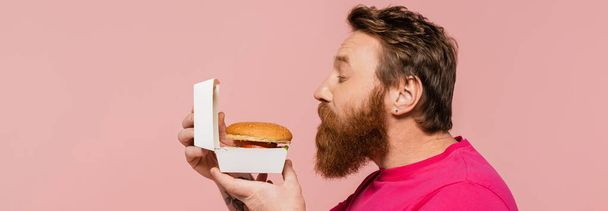 side view of bearded man smelling delicious hamburger isolated on pink, banner - 写真・画像