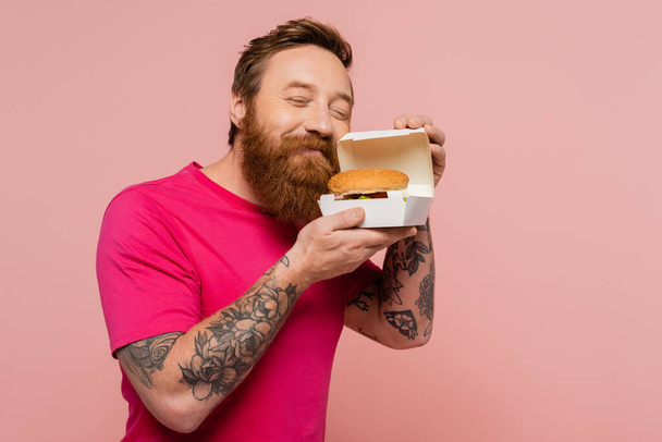 joyful tattooed man with closed eyes smelling tasty burger in carton box isolated on pink - 写真・画像