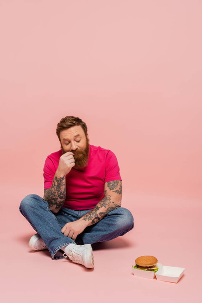 full length of thoughtful man in jeans looking at carton pack with burger while sitting with crossed legs on pink background - Photo, Image