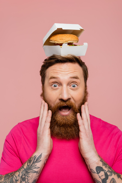 astonished man with hamburger in carton pack on head touching beard and looking at camera isolated on pink - Photo, Image