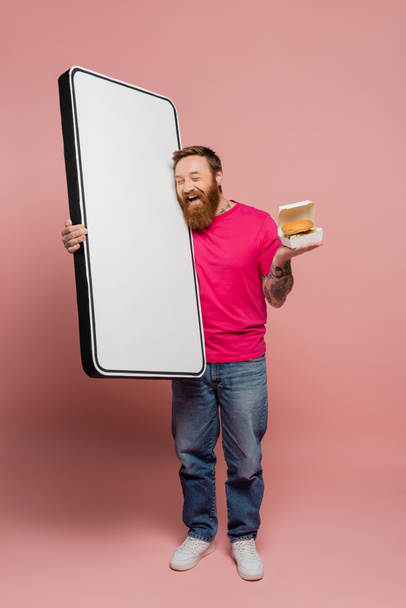 bearded man holding hamburger and huge template of mobile phone while laughing with closed eyes on pink background - Photo, Image
