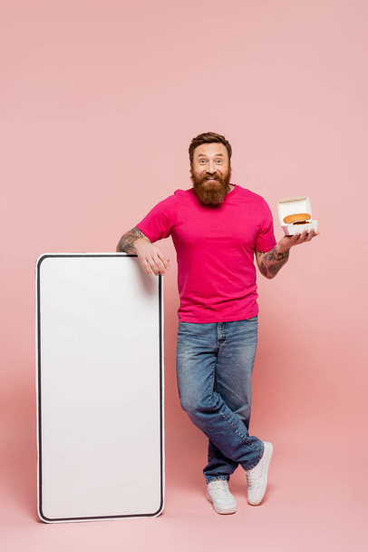 full length of cheerful man in jeans standing with hamburger near big template of smartphone on pink background - Photo, image