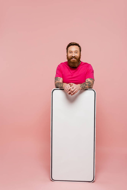 homem barbudo e tatuado em magenta t-shirt olhando para a câmera perto de modelo de telefone enorme no fundo rosa - Foto, Imagem