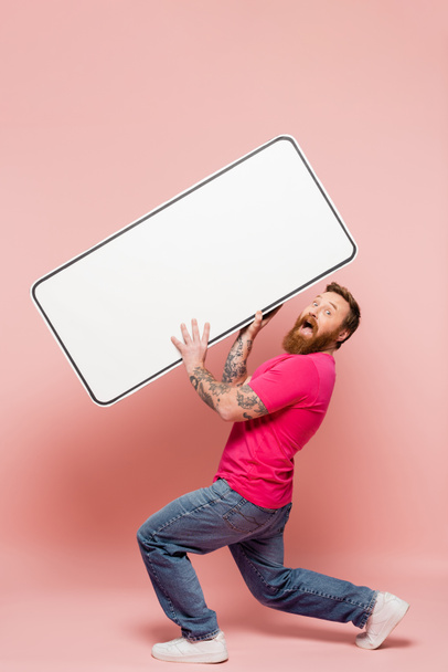 full length of astonished man looking at camera while holding big phone template on pink background - Foto, Bild