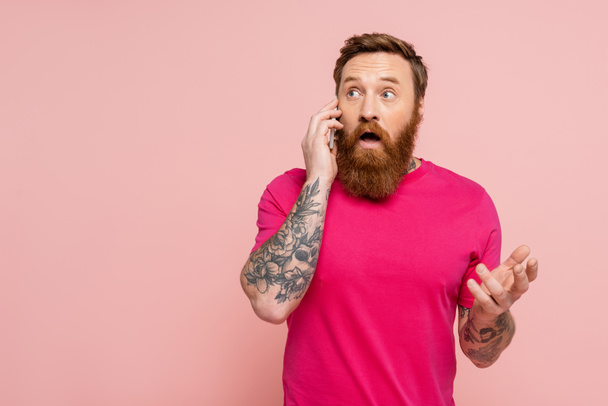 verraste man met baard in magenta t-shirt weg te kijken tijdens het praten op smartphone geïsoleerd op roze - Foto, afbeelding